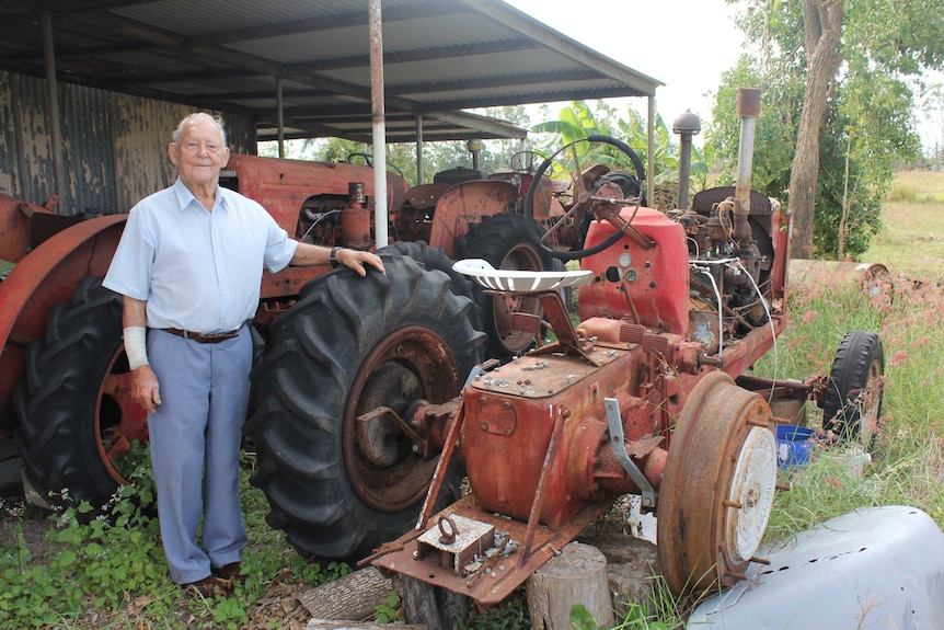 A man stands beside the large wheel of a tractor in a shed filled with other tractors