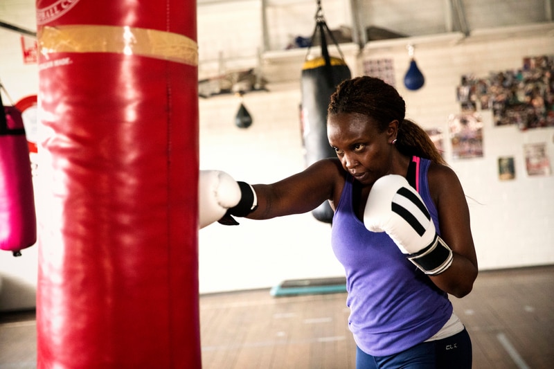 Joys Njambi wearing white boxing gloves and hitting a red punching bag in a gym.