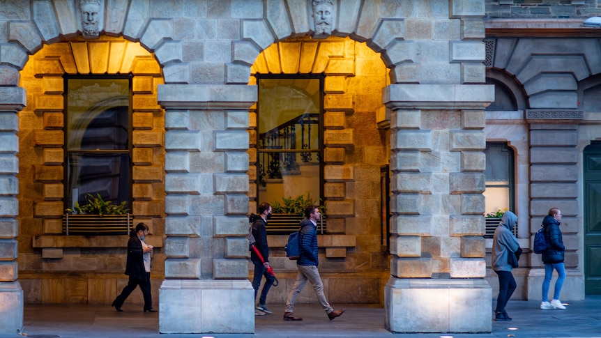 People walking on a footpath with arches of a 19th century stone building