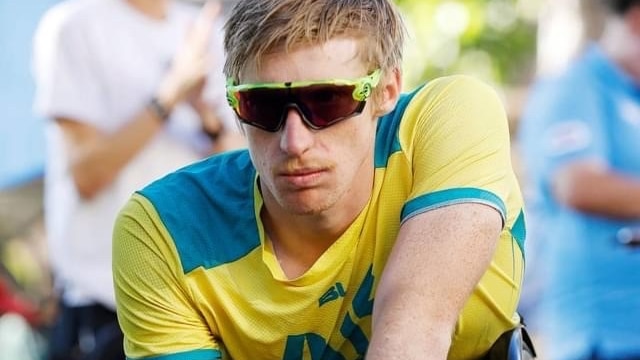 A young man in the green and gold and wrap around shades in a racing wheelchair.