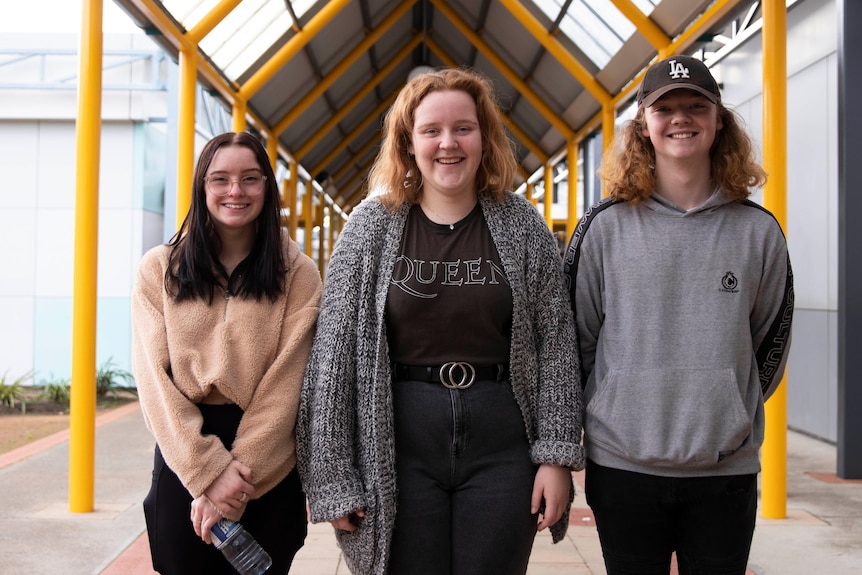 Three teens stand in a line and smile outside a schoo building.