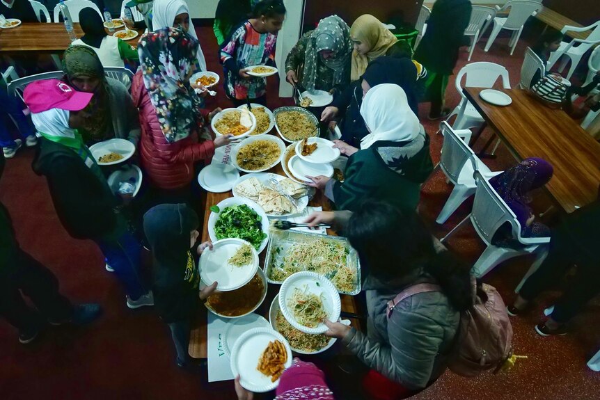 A group of children and woman crowd around a table laden with dishes of hot noodles and rice.