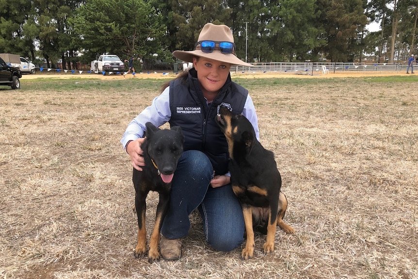 A woman kneels down to hug two kelpies in a paddock.