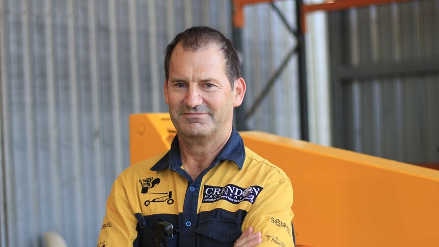 A man in a yellow work short stands in front of yellow machinery and a corrugated iron fence