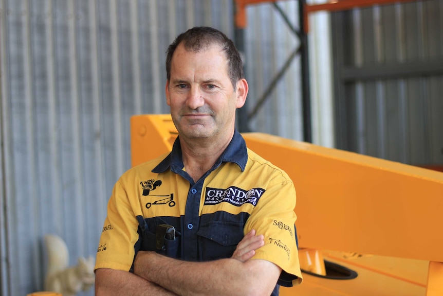 A man in a yellow work short stands in front of yellow machinery and a corrugated iron fence