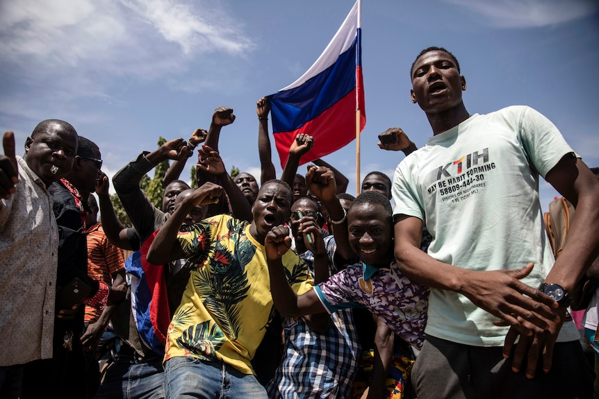 A group of men is pictured sticking their fists in the air while seemingly shouting/chanting. Behind them a Russian flag.