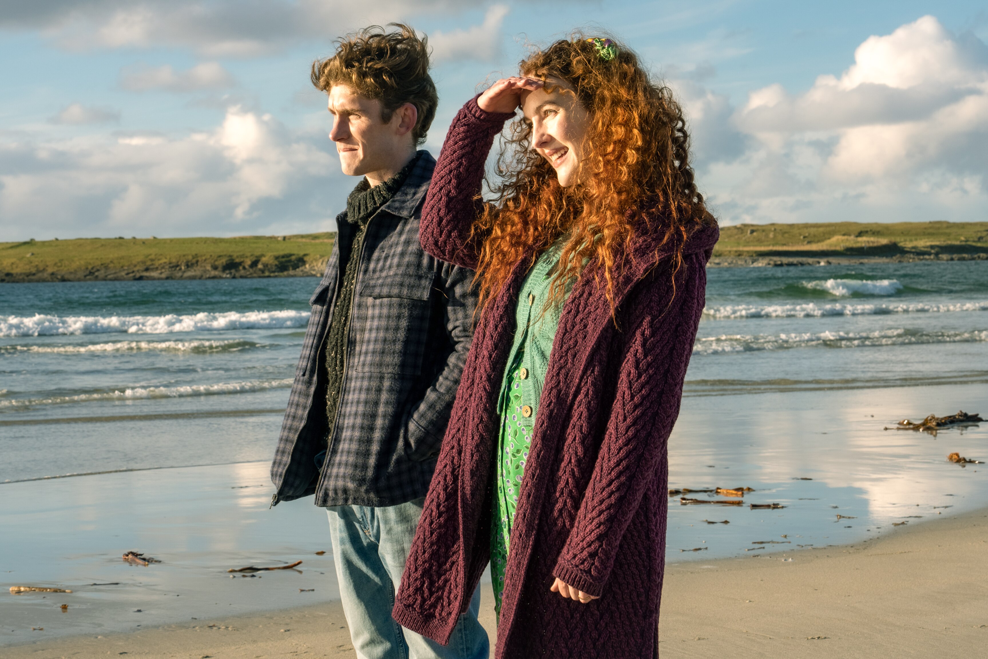 Fionn and Ann look to the left, with Ann holding her hand up to her face to block out the sun while they stand on the beach.