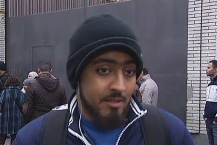Houcine is a young man with a beard and wearing a beanie, jip-up jumper and backpack. He stands in front of a small crowd.