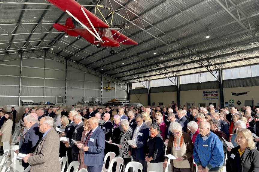 People standing in an airport hangar with a vintage red plane above.