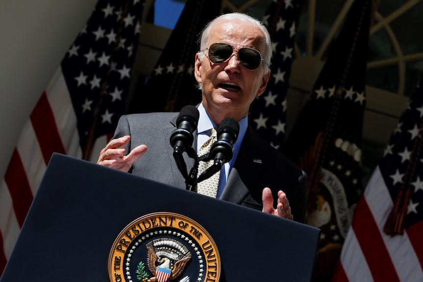 Joe Biden speaks at a podium. He wears sunglasses and a suit. There are US flags behind him.