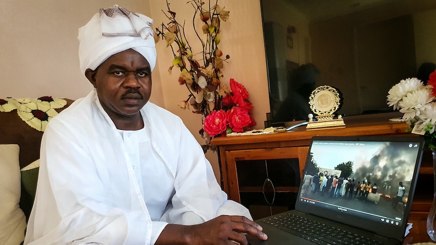 Un african sumbru de vârstă mijlocie, într-un costum tradițional alb, urmărește un ecran video cu scene violente africane