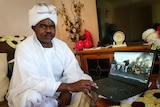 A sombre middle-aged African man in white traditional suit watches a video monitor of African violent scenes