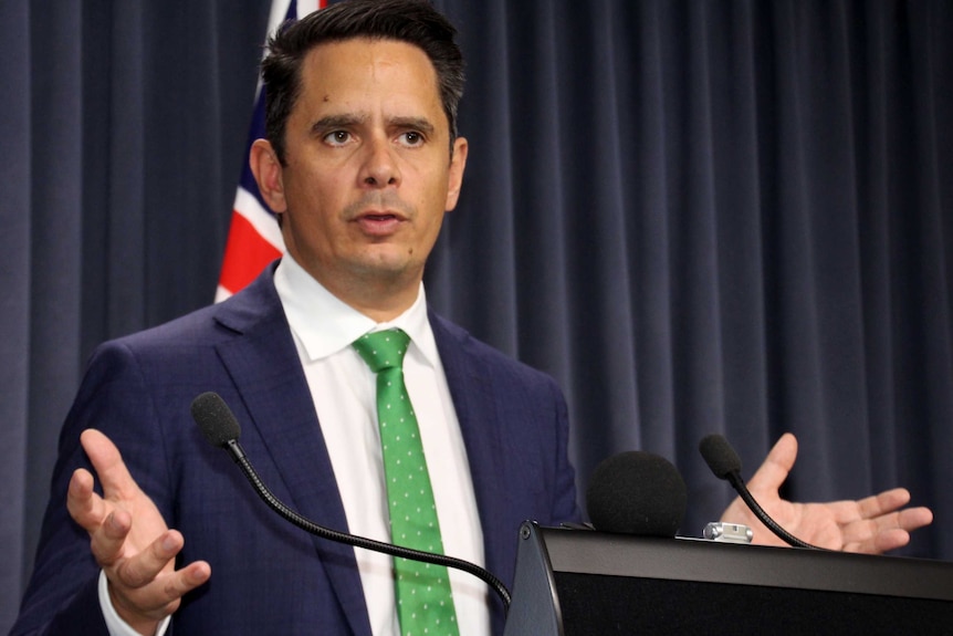 WA Treasurer Ben Wyatt stands at a microphone with an Australian flag in the background.