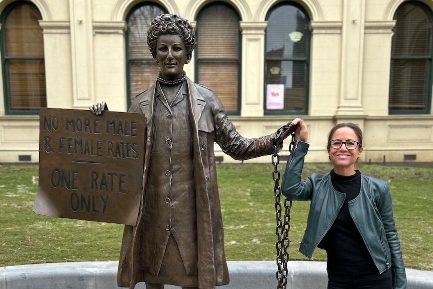 Woman holding hand of bronze statue of woman with sign saying 'no more male and female rates, one rate only' 