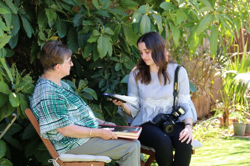 A young female reporter holds a notepad and camera while sitting in a garden chatting with a woman