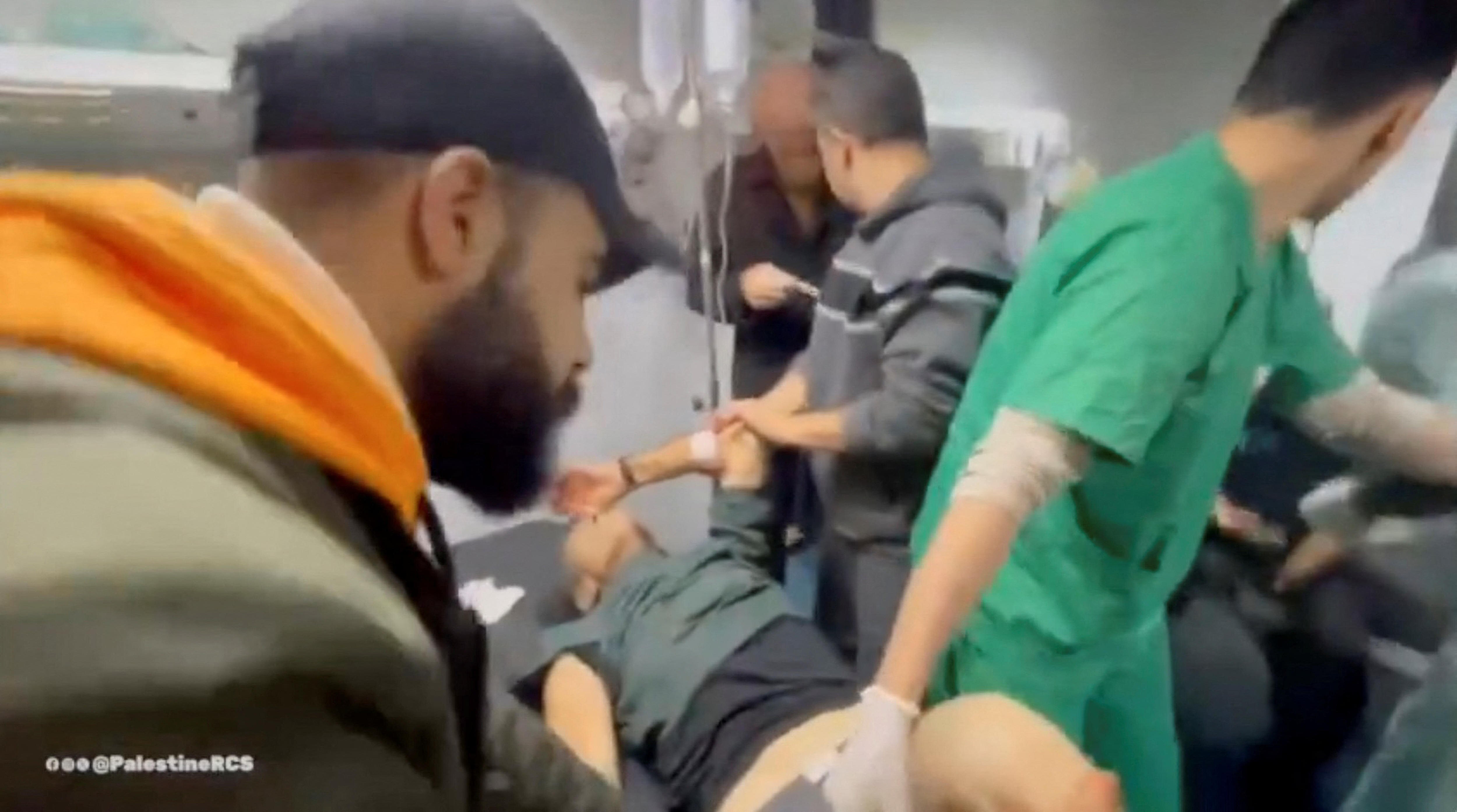 A screenshot from a video showing a man lying on a hospital bed while being treated by a male nurse. Other men stand nearby.