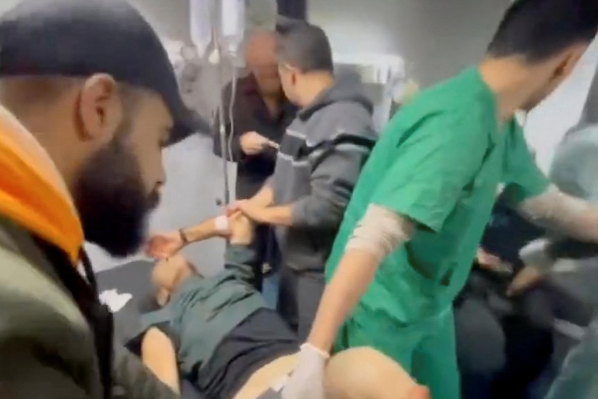 A screenshot from a video showing a man lying on a hospital bed while being treated by a male nurse. Other men stand nearby.