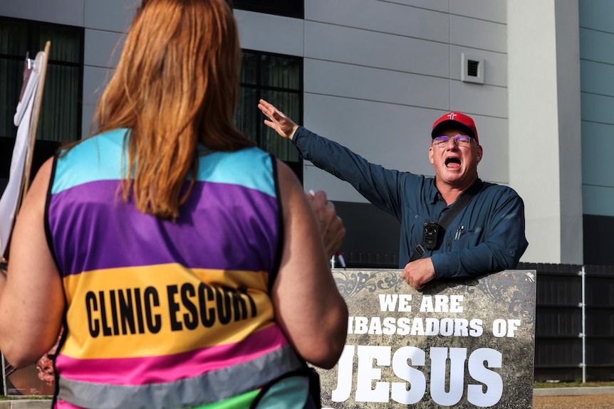 A man with a sign saying WE ARE THE AMBASSADORS OF JESUS yells and gestures at a woman with CLINIC ESCORT on her shirt