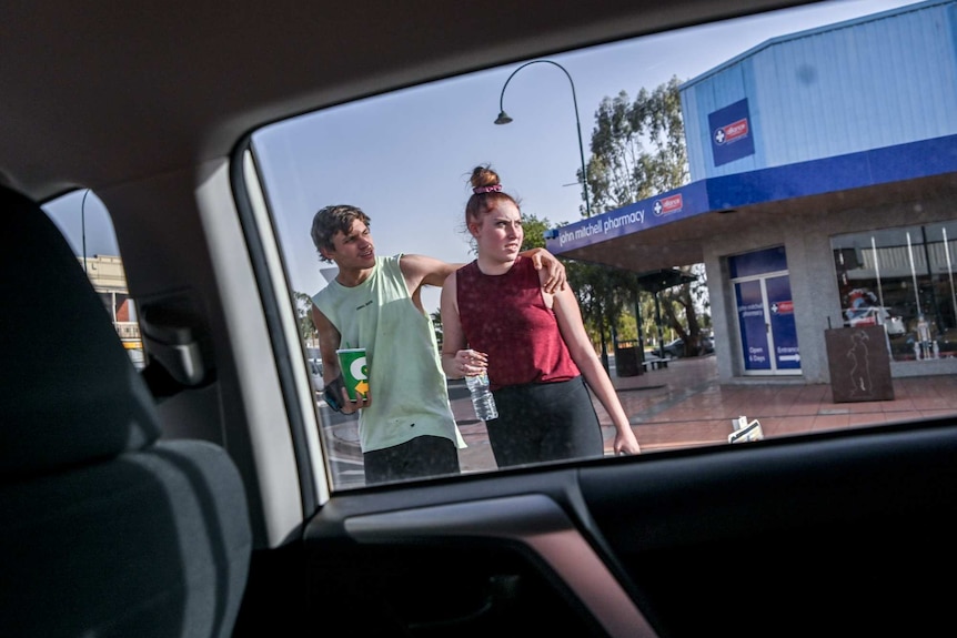 Two Cobar residents, one with an arm around the other, in the street seen out the window of a car.