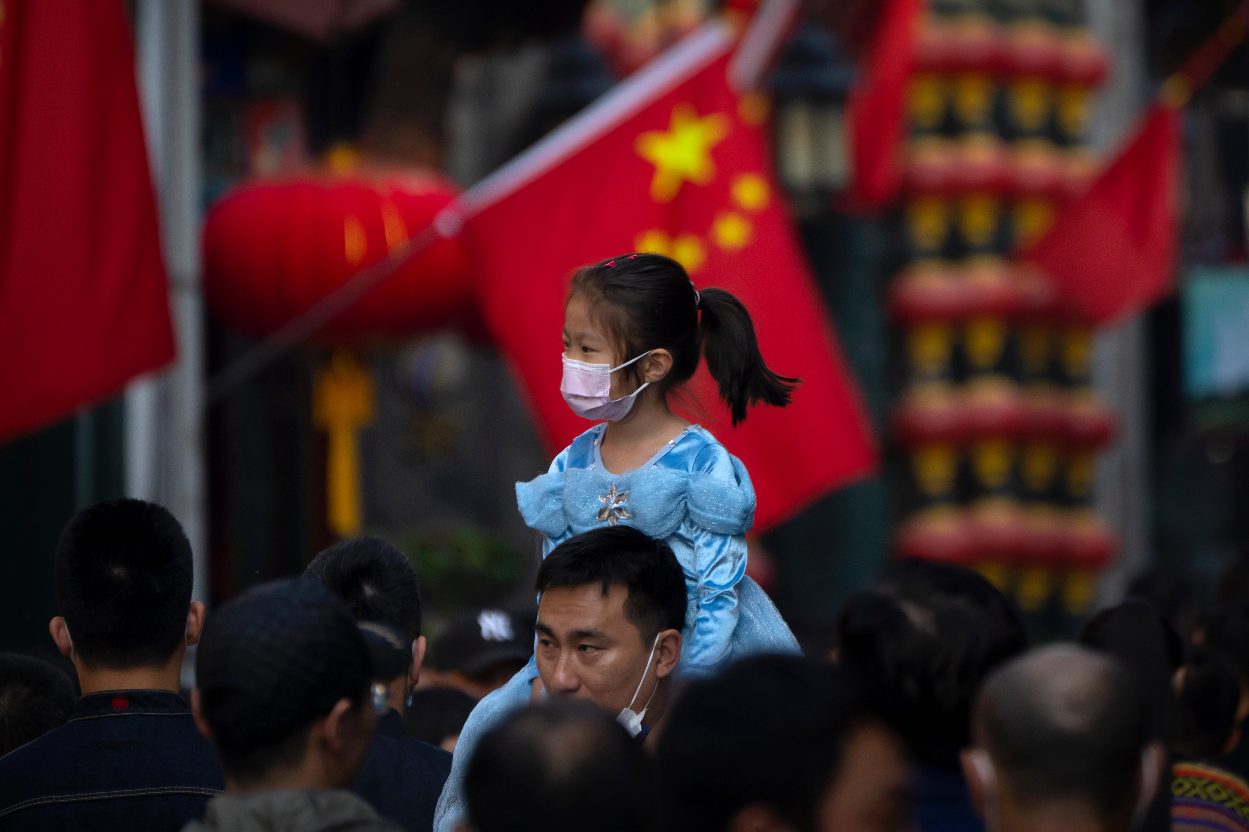 一名戴着口罩的女孩骑在一名男子的肩膀上，他们走在北京的一条旅游购物街上。