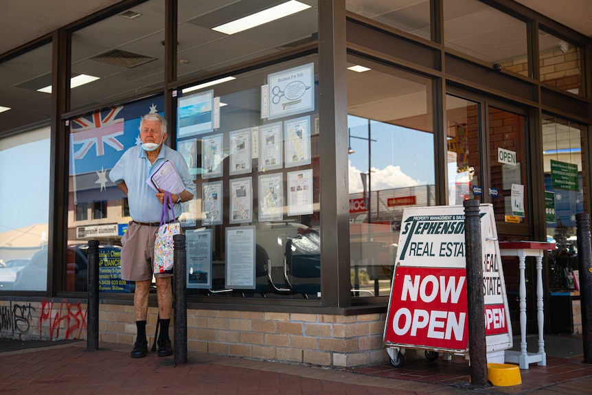An older man stands outside a real estate displaying an Australian flag in the window.