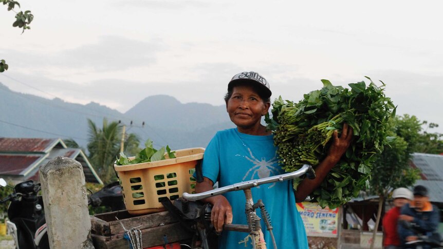 Mid-shot of Poso woman, resting on a bike and holding green producer, with mountains in the background.