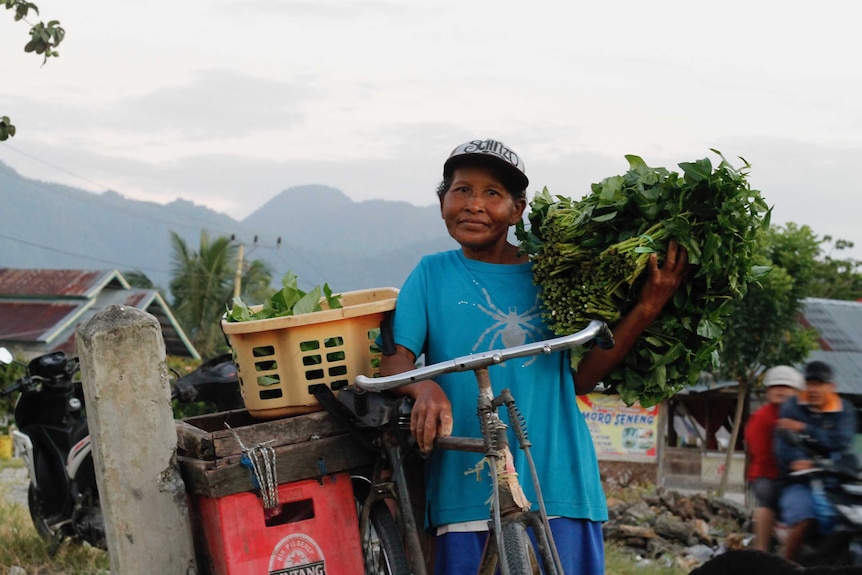 Mid-shot of Poso woman, resting on a bike and holding green producer, with mountains in the background.