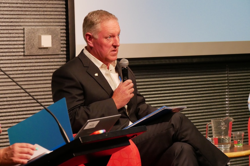 A man in a suit sitting in a chair, holding a microphone to his mouth