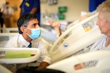 A middle-aged man of Indian descent wears a mask as he kneels to speak at the bedside of an elderly white woman in a hospital.