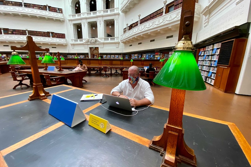 A man in a mask sits at a desk in a room surrounded by bookshelves.