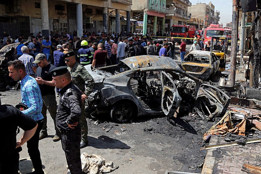 The shell of a blackened and burnt out car is surrounded by officers in Baghdad