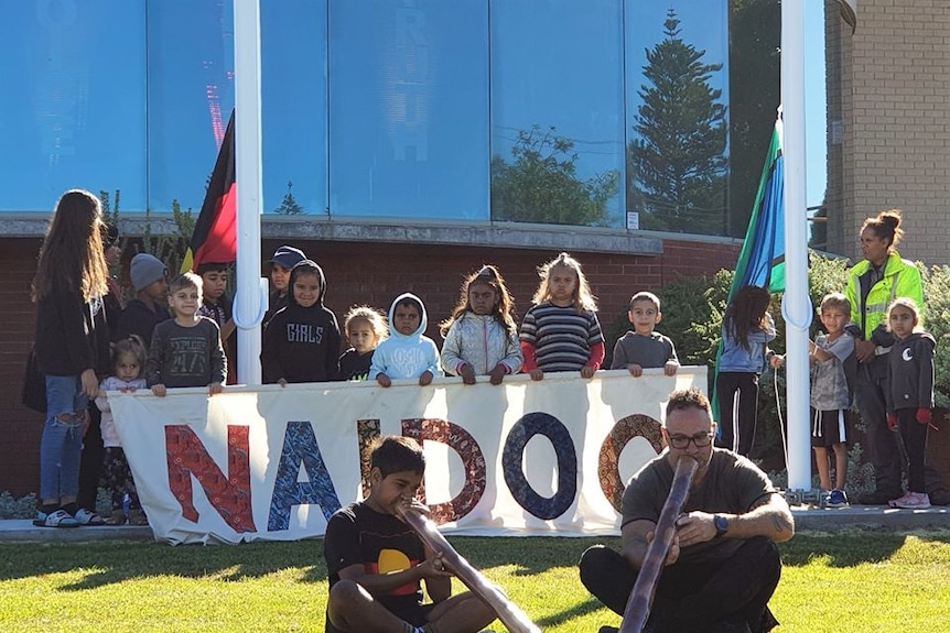 Two men playing a didgeridoo in front of children and the Aboriginal flag.