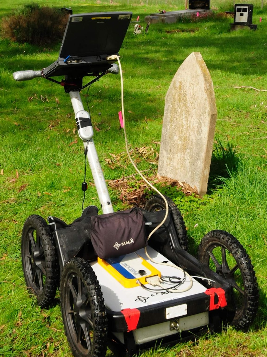 A lawnmower-type device with a laptop on the handlebars sits in a lush bushland cemetery.