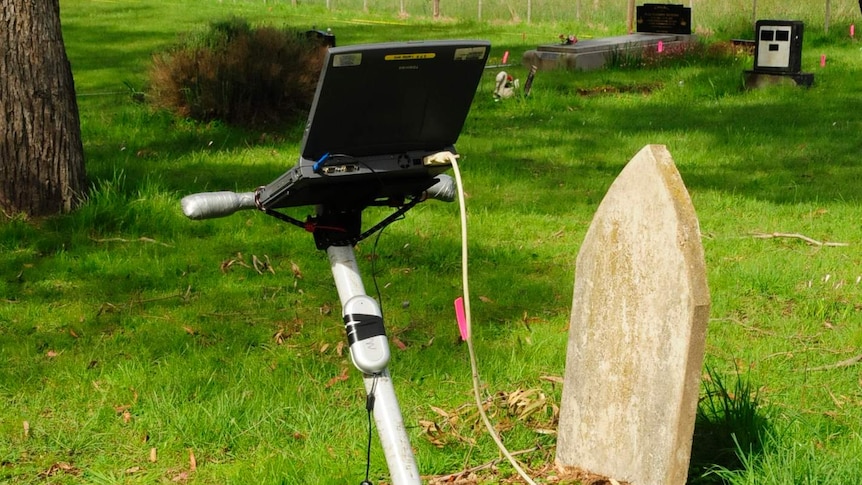 A lawnmower-type device with a laptop on the handlebars sits in a lush bushland cemetery.