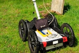 A lawnmower-type device with a laptop on the handlebars sits in a lush bushland cemetery.