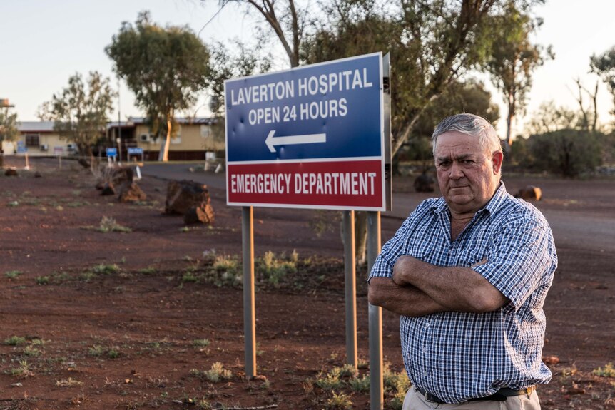 man with arms crossed stands in front of sign pointing toward hospital entrance