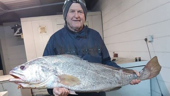 Greg Sichter wears a beanie and fishing clothes, holding a large Black Jewfish in his garage.