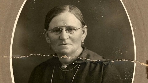 A woman is sitting for her photo. It is in black and white and she is wearing glasses, a necklace and has her hair pulled back.