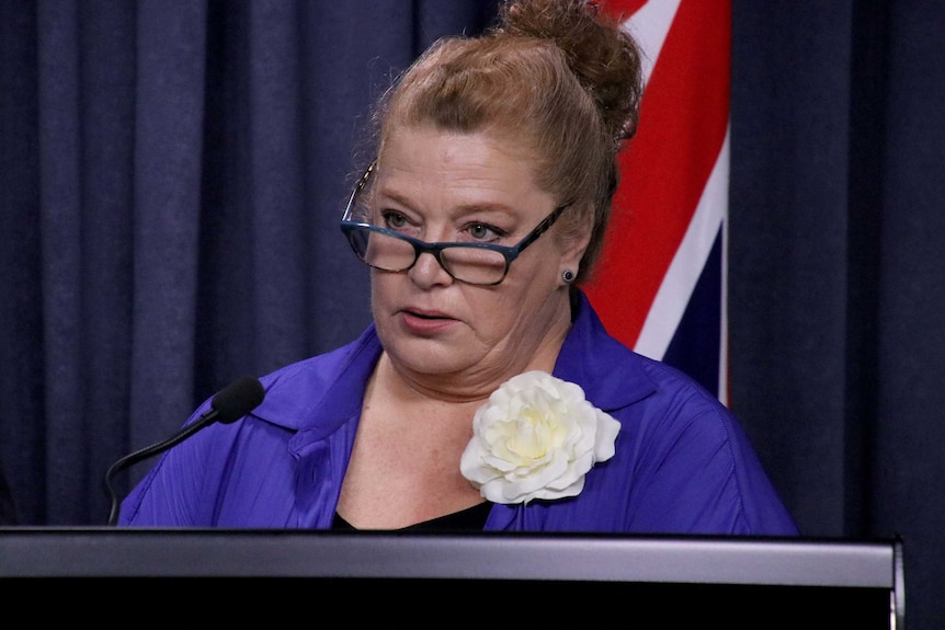 A serious looking woman standing at a lectern with an Australian flag in the background.