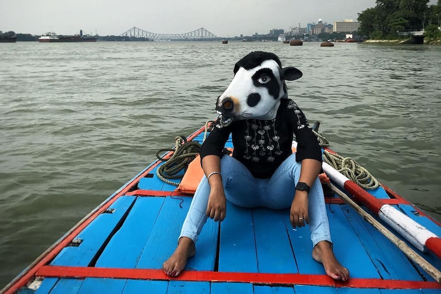 A woman wears a cow mask while sitting on a boat on a river in Kolkata.