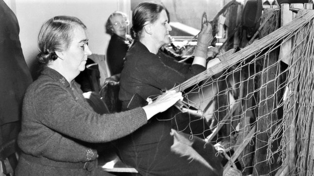 A black and white photograph of two older women sitting assembling a large net.