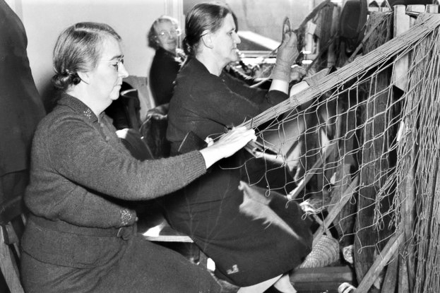 A black and white photograph of two older women sitting assembling a large net.