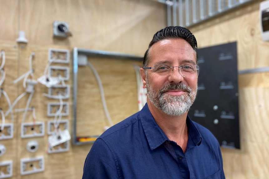 A man with a beard and wearing a blue shirt stands in front of an electrical board.