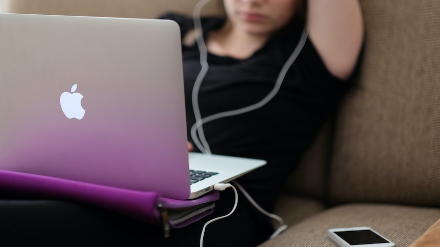 A teenage girl in a black tshirt reclines on a sofa with a macbook on her lap and an iphone beside her