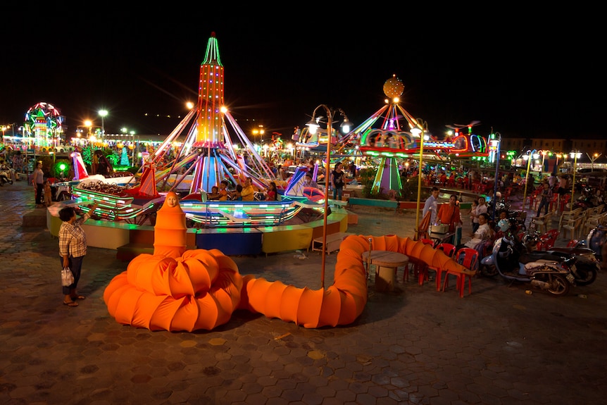 Woman in bright orange caterpillar-like chador costume standing in fairground at night.