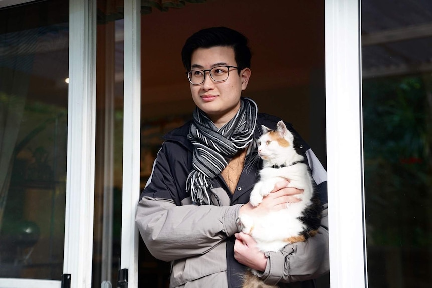 A picture of a young man in scarf and glasses holding a cat and looking out a window.