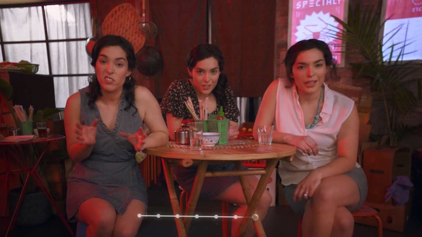 Three women, seated in a restaurant, talking directly at camera.