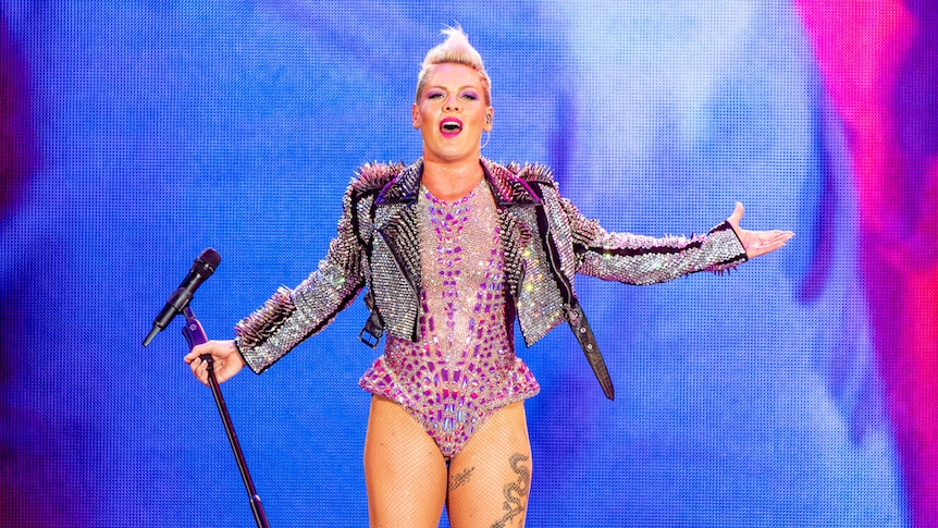 Pink stands on stage holding a mic stand as she glitters in a pink bodysuit and studded black leather jacket.