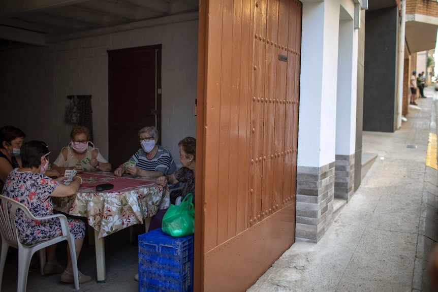 Five women wearing face masks play cards inside a house.