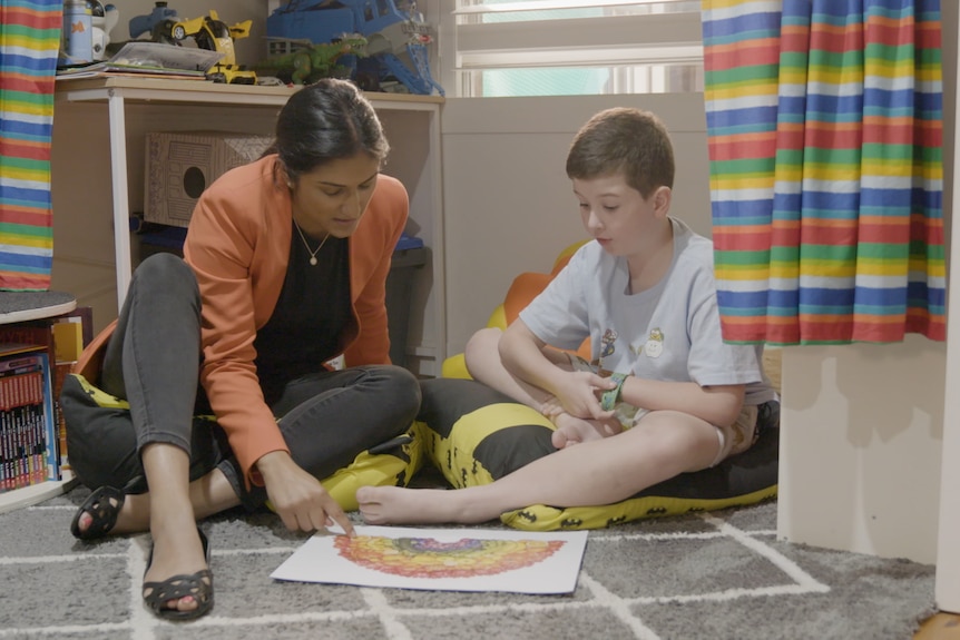 A female doctor sits with a little boy in a cubby, looking at a poster with fruit and vegetables on it.